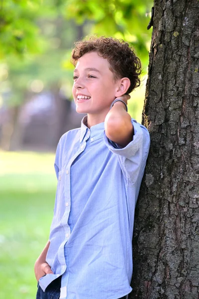 Молодий щасливий хлопчик розслабляється в літньому парку, спираючись на стовбур дерева. Позитивний юнак насолоджується літнім часом на відкритому повітрі. Концепція добробуту дітей — стокове фото