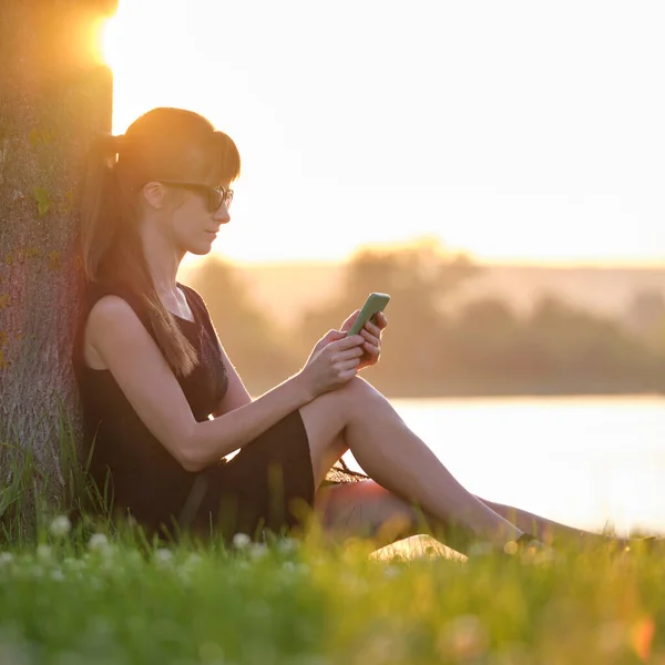 Молодая женщина отдыхает на лужайке возле озера, просматривая свой мобильный телефон на открытом воздухе в теплый летний вечер. Концепция связи и мобильной связи — стоковое фото