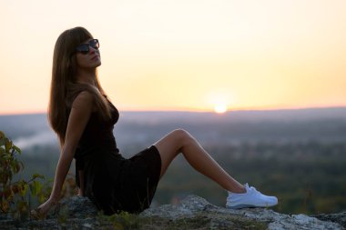 Siyah kısa yaz elbisesi içinde genç ve güzel bir kadın gün batımında bir kayanın üzerinde oturup dinleniyor. Modaya uygun bir kadın doğada sıcak bir akşamın tadını çıkarıyor.