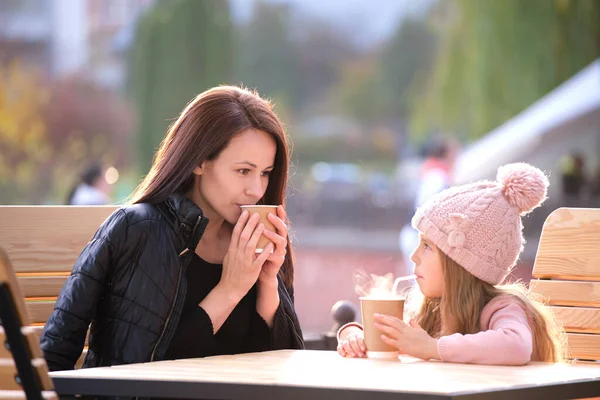 젊은 어머니와 딸은 화창 한 가을에 뜨거운 음료를 마시며 거리 카페에서 즐거운 시간을 보내고 있다. 가족 관계 에서의 행복 — 스톡 사진