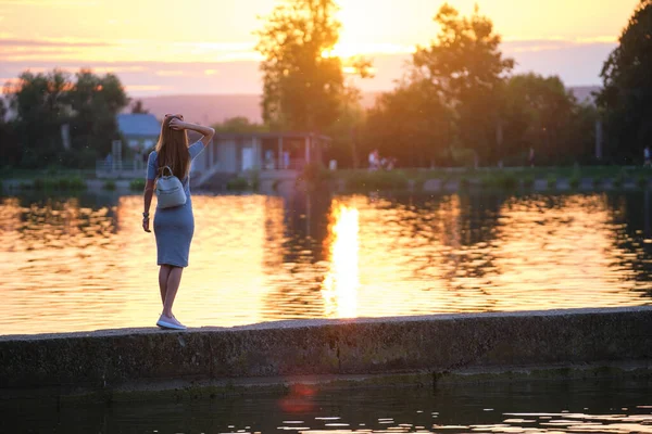 Одинокая молодая женщина, стоящая одна на берегу озера и наслаждающаяся теплым вечером. Благополучие и отдых в природе — стоковое фото