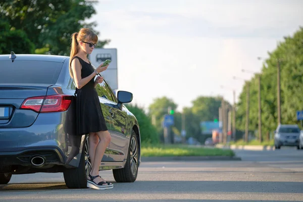 Стильная женщина-водитель, стоящая у своего автомобиля и говорящая по мобильному телефону на городской улице летом — стоковое фото