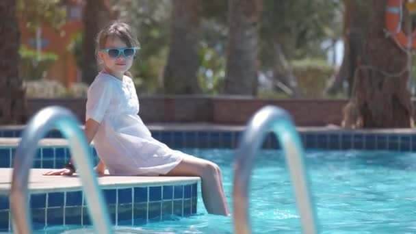 Joyeux enfant fille en robe blanche relaxant sur le côté de la piscine le jour ensoleillé d'été — Video