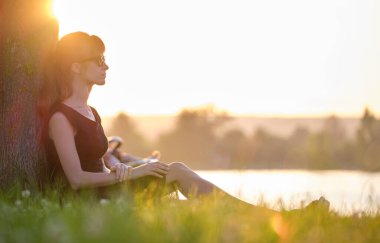 Yalnız bir kadın, sıcak bir akşamda yeşil çimenlerde tek başına oturmuş göl kıyısındaki ağaç gövdesine yaslanıyor. Yalnızlık ve doğa konseptinde rahatlama.