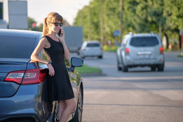 Молодая женщина-водитель, стоящая рядом с машиной и разговаривающая по мобильному телефону на городской улице летом — стоковое фото