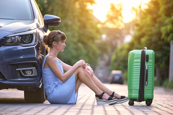 Joven mujer cansada con la maleta sentada cerca de su coche esperando a alguien. Concepto de viajes y vacaciones — Foto de Stock