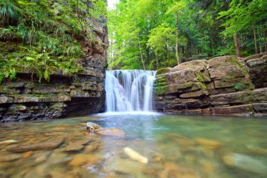 Yazın yağmur ormanlarında kayalık kayalıklardan dökülen beyaz köpüklü suyla dağ nehrindeki muhteşem şelale manzarası.