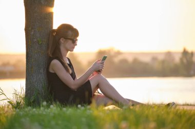 Genç bir kadın göl kenarındaki çimenlerde rahat rahat cep telefonuna bakıyor ılık bir yaz akşamında. İletişim ve mobil bağlantı kavramı