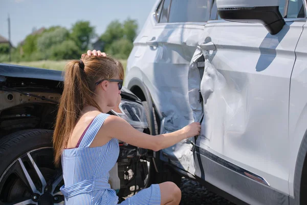 Sad young woman driver sitting near her smashed car looking shocked on crashed vehicles in road accident — Stock Photo, Image