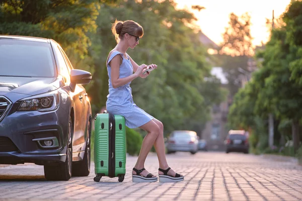 Young tired woman with suitcase sitting beside car waiting for someone. Travelling and vacations concept