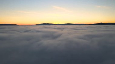 Beyaz yoğun bulutlar ve ufuktaki uzak dağların üzerinde canlı sarı gündoğumunun havadan görüntüsü.