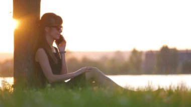 Yaz parkında gölün kıyısında oturan genç bir kadın sıcak bir akşamda cep telefonuyla dışarıda konuşuyor. İletişim ve mobil bağlantı kavramı