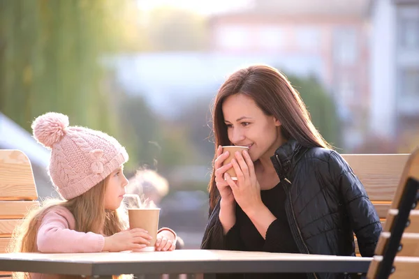 젊은 어머니와 딸은 화창 한 가을에 뜨거운 음료를 마시며 거리 카페에서 즐거운 시간을 보내고 있다. 가족 관계 에서의 행복 — 스톡 사진