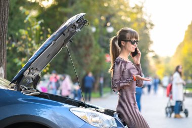 Kızgın kadın sürücü, bozuk bir arabanın yanında duran yardım görevlisiyle cep telefonuyla konuşuyor.