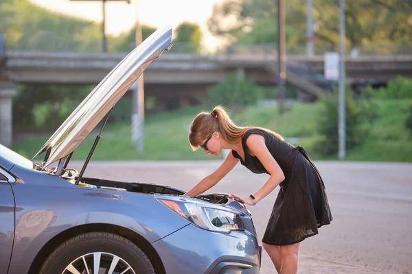 Mujer indefensa de pie cerca de su coche con el capó abierto inspeccionando el motor roto. Conductora joven que tiene problemas con el vehículo — Foto de Stock