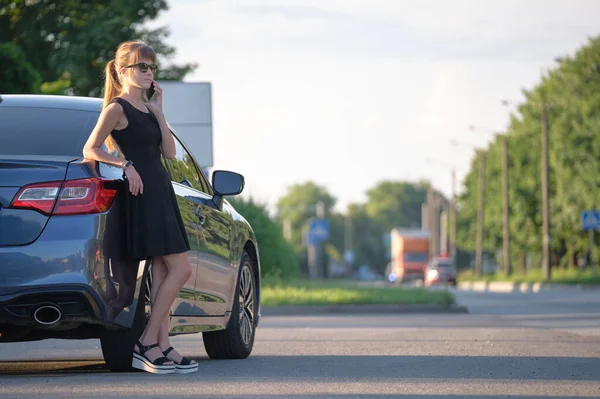 Стильная женщина-водитель, стоящая у своего автомобиля и говорящая по мобильному телефону на городской улице летом — стоковое фото