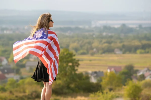 Młoda szczęśliwa amerykańska kobieta z długimi włosami machająca na wietrze flagą USA narodowa na jej sholders relaks na świeżym powietrzu ciesząc się ciepły letni dzień — Zdjęcie stockowe