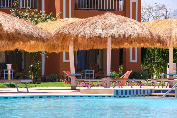 Sillas de cubierta vacías bajo sombrillas de paja en el lado de la piscina en el complejo tropical. Vacaciones de verano y concepto de escapada —  Fotos de Stock