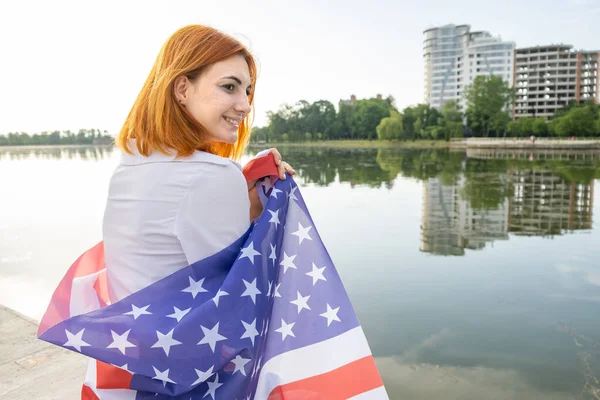 Szczęśliwy uśmiechnięty młoda kobieta z flagą narodową USA na ramionach z wysokich budynków miejskich w tle świętując dzień niepodległości Stanów Zjednoczonych — Zdjęcie stockowe