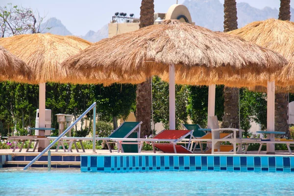 Sillas de cubierta vacías bajo sombrillas de paja en el lado de la piscina en el complejo tropical. Vacaciones de verano y concepto de escapada — Foto de Stock