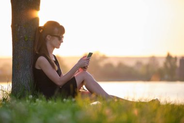 Genç bir kadın, göl kıyısındaki çimlerin üzerinde oturmuş sıcak bir yaz akşamında cep telefonunu karıştırıyor. İletişim ve mobil bağlantı kavramı