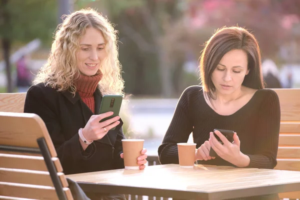 Two distracted female friends browsing their mobile phones while sitting together at city street cafe. Girlfriends ignoring each other while staring in gadgets. Addiction from social media concept — стоковое фото