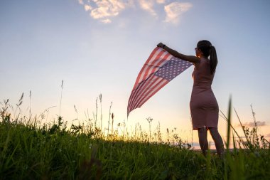 Gün batımında ABD bayrağıyla dışarıda poz veren mutlu genç bir kadının arka görüntüsü. Amerika 'nın bağımsızlık gününü kutlayan pozitif kız. Uluslararası Demokrasi Günü.