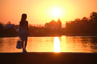 Sıcak bir akşamda göl kenarında rahatlayan günlük kıyafetli genç bir kadın. Yaz tatilleri ve seyahat konsepti.