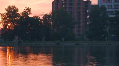 Göl suyuna yansıyan karanlık bina silüetiyle Lakeside manzarası ve parlak günbatımında toprak üzerinde yürüyen yayalar..