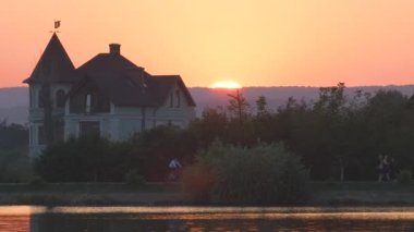 Göl suyunun önündeki banliyö evlerinin karanlık silueti ve gün batımında sette yürüyen yayalar..
