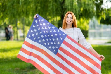Mutlu kızıl saçlı kadın yaz parkında Amerikan bayrağıyla poz veriyor. Açık havada afişi olan pozitif kız..