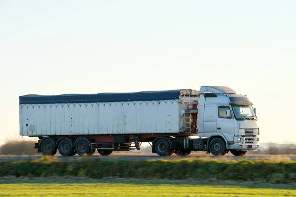 Semi-truck with tipping cargo trailer transporting sand from quarry driving on highway hauling goods in evening. Delivery transportation and logistics concept.