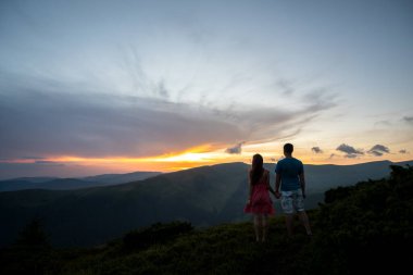 Genç yürüyüşçü çiftin çimenli dağ tepesinde el ele tutuşup günbatımı manzarasının ve dramatik akşam gökyüzünün tadını çıkarışının arka görüntüsü..