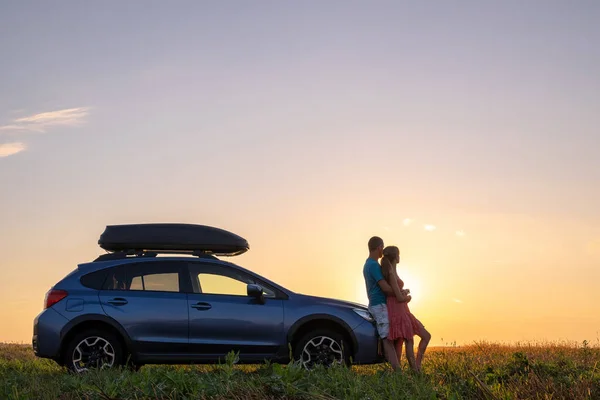 Happy couple standing near their car at sunset. Young man and woman enjoying time together travelling by vehicle.