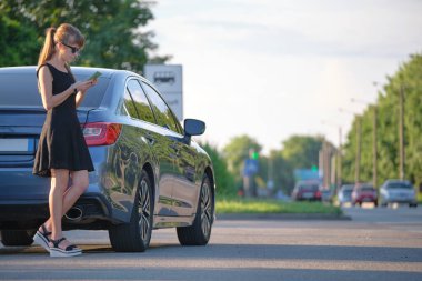 Arabasının yanında duran genç bir kadın sürücü yazın bir şehir caddesinde cep telefonuyla konuşuyor..