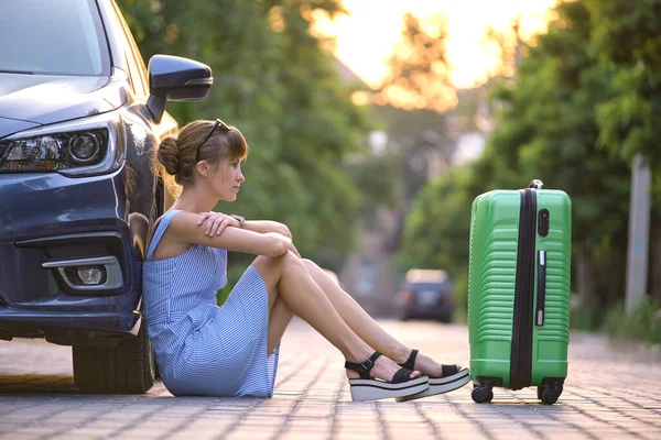 Young tired woman with suitcase sitting beside car waiting for someone. Travelling and vacations concept.