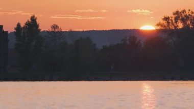 Göl suyuna yansıyan karanlık park ağaçlarının gölgeli manzarası ve gün batımında parkın tepesinde yürüyen yayalar..