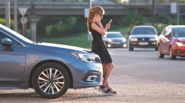 Arabasının yanında duran genç bir kadın sürücü yazın bir şehir caddesinde cep telefonuyla konuşuyor..