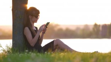Genç bir kadın göl kenarındaki çimenlerde rahat rahat cep telefonuna bakıyor ılık bir yaz akşamında. İletişim ve mobil bağlantı kavramı.