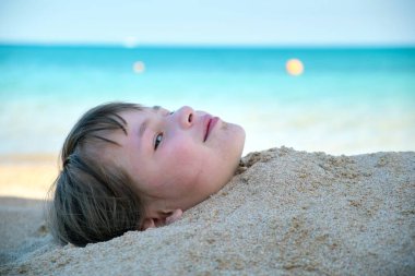 Young smiling child girl lying down covered with white sand on tropic beach on blue sky and ocean water background.