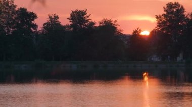 Göl suyunda kırılan park ağaçlarından oluşan karanlık bir siluet ve gün batımında parkın tepesinde yürüyen yayalar ile Lakeside manzarası.