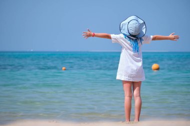 Child girl in white dress and hat standing barefooted with wide spread hands on beach enjoying tropic vacations looking at ocean water.