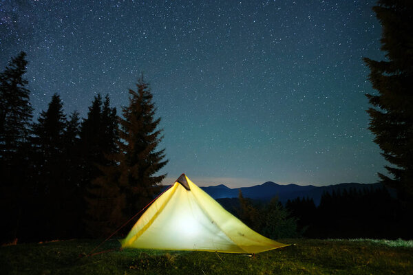 Bright illuminated tourist tent glowing on camping site in dark mountains under night sky with sparkling stars. Active lifestyle concept.