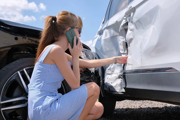 Stressed driver talking on sellphone on roadside near her smashed vehicle calling for emergency service help after car accident. Road safety and insurance concept.