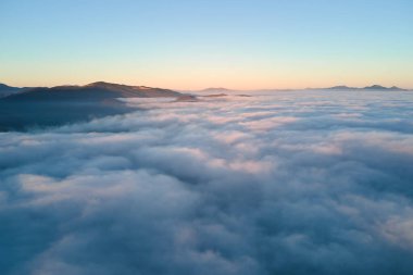 Ufuktaki dağ tepelerinin uzak karanlık siluetleri ile beyaz yoğun sis üzerinde renkli gün doğumunun havadan görüntüsü.