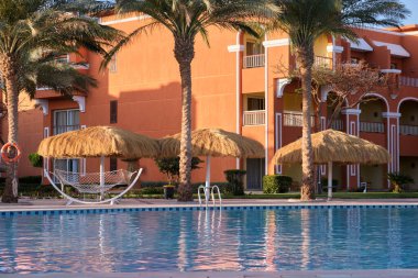 Boş güverte sandalyeleri, tropik tatil köylerinde havuz kenarında saman gölgesi şemsiyelerinin altında. Yaz tatilleri ve kaçış konsepti.