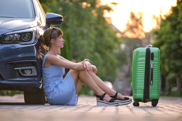 Young tired woman with suitcase sitting near her car waiting for someone. Travel and vacations concept.