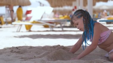 Happy child girl in swimsuit playing with sand under umbrella shadow during summer tropical vacations.