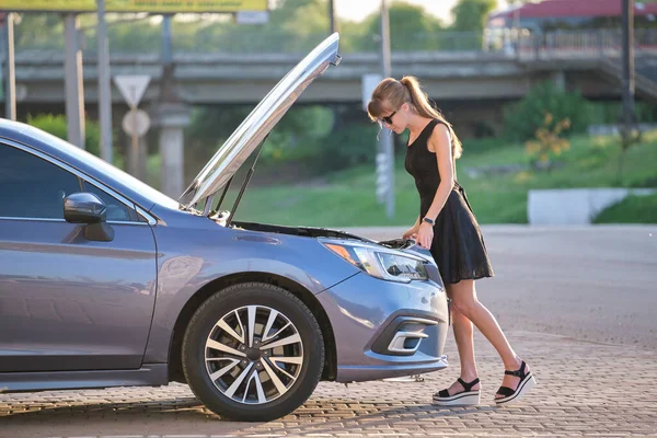 Kafası karışmış bir kadın sürücü arabasının yanındaki caddede durmuş kaputu patlamış ve bozuk motora bakıyor..