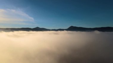 Geniş dağ vadisindeki beyaz kabarık bulutların üzerinde ilerleyen hava görüntüsü.